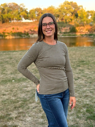 A woman wearing a long-sleeve crewneck shirt with thin horizontal stripes in olive green and beige, paired with blue jeans, standing outdoors in front of a grassy field and a lake.