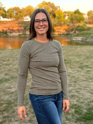 A woman wearing a long-sleeve crewneck shirt with thin horizontal brown and beige stripes, paired with blue jeans, standing outdoors in front of a grassy field and a river.