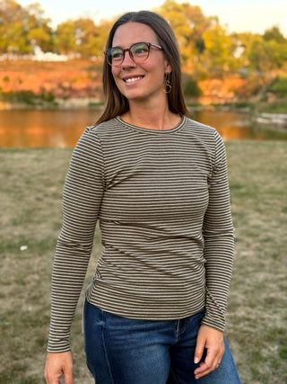 A woman wearing a long-sleeve crewneck shirt with thin horizontal brown and beige stripes, paired with blue jeans, standing outdoors in front of a grassy field and a lake at sunset.