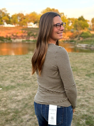 A woman with long brown hair and glasses, wearing a beige and brown horizontally striped long-sleeve top and blue jeans, standing outdoors in a grassy field with a lake and autumn trees in the background.