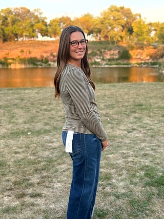 A woman with long brown hair and glasses stands outdoors in a grassy field near a lake, wearing a beige and dark gray striped long-sleeve top and blue jeans.