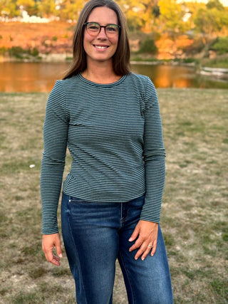 A woman wearing a dark teal and navy striped long-sleeve shirt with blue jeans, standing outdoors in front of a grassy field and a lake.