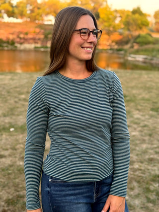 A woman wearing a dark teal and white striped long-sleeve shirt with blue jeans, standing outdoors in front of a park lake at sunset.