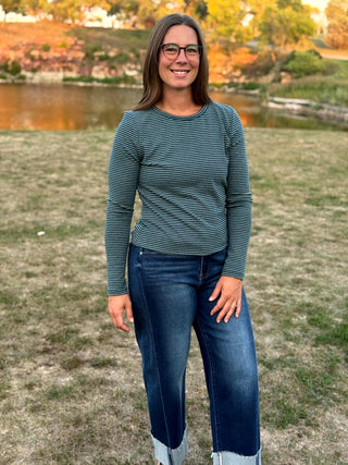 A woman wearing a dark green and gray striped long-sleeve top and dark blue jeans stands outdoors on a grassy lawn with a lake and trees in the background.