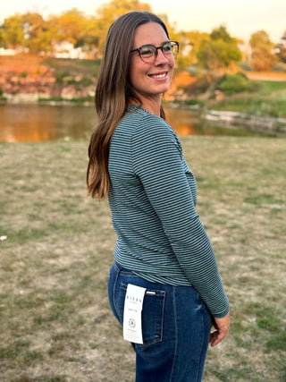 A woman wearing a slate blue and gray striped long-sleeve top and dark blue jeans, standing outdoors in a grassy field with trees and a pond in the background. A white paper tag is attached to the back pocket of her jeans.