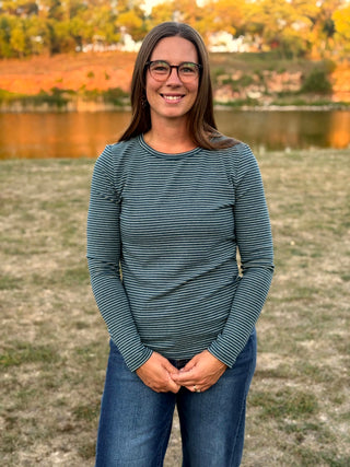 A woman with glasses stands outdoors in front of a lake at sunset, wearing a dark teal and gray striped long-sleeve shirt and blue jeans.