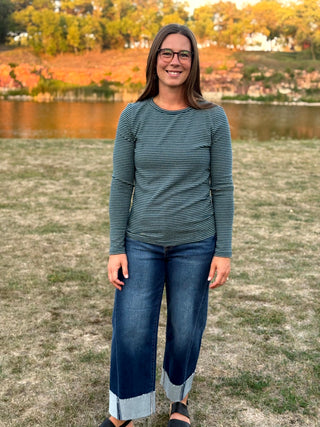 A woman wearing a dark green and gray striped long-sleeve top and blue jeans with rolled cuffs, standing outdoors in front of a grassy field and a lake.