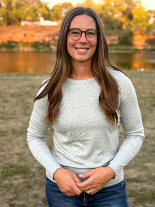 A smiling woman with long brown hair and glasses stands outdoors in front of a lake, wearing a light gray long-sleeve crewneck shirt and blue jeans.