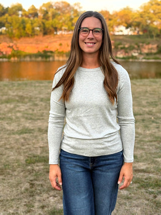 A woman with long brown hair and glasses stands outdoors in front of a lake and autumn trees, wearing a light heather gray long-sleeve crewneck shirt and blue jeans.