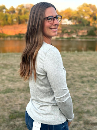 A woman with long brown hair and glasses, wearing a light gray long-sleeve shirt and blue jeans, standing outdoors in a grassy field near a lake at sunset.