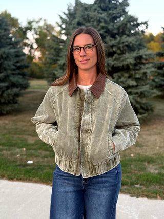 A woman wearing a light olive green corduroy jacket with a brown collar, blue jeans, and glasses, standing outdoors in front of trees.