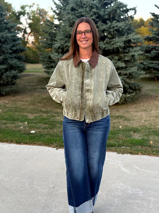 A woman wearing a light olive green corduroy bomber jacket with a brown collar and blue jeans, standing outdoors on a paved path with trees in the background.