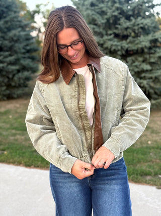 A woman wearing a light sage green corduroy jacket with a brown corduroy collar and front zipper, styled over a cream t-shirt and blue jeans, standing outdoors in front of trees.