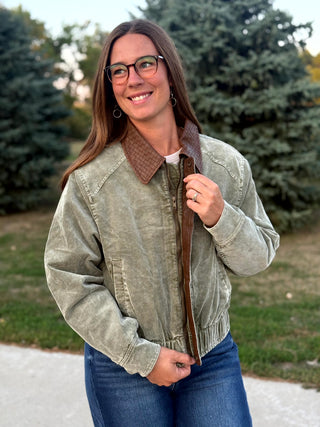 A woman wearing a light olive green corduroy jacket with a brown collar and zipper trim, paired with blue jeans, standing outdoors in front of trees.