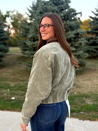 A woman with long brown hair and glasses, wearing a sage green corduroy jacket and blue jeans, photographed outdoors in a park setting with trees in the background.