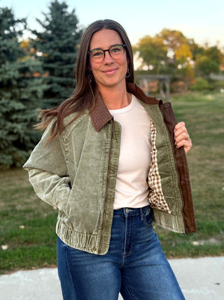 A woman wearing a light olive green corduroy jacket with brown collar and lining, styled over a cream t-shirt and blue jeans, standing outdoors in a park setting.