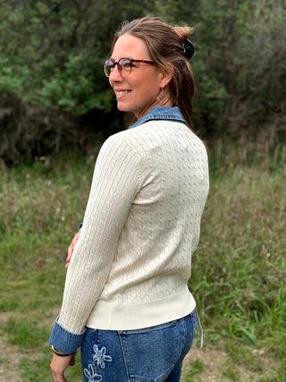 A lady wearing a ivory cable knit sweater with denim trim on the collar and sleeves.