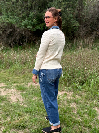 A person standing outdoors in a grassy field, shown from the back wearing blue jeans with a cream sweater and black shoes. A paper tag is attached to the back pocket of the jeans.