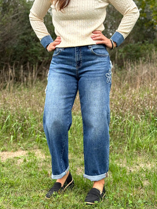 A person standing outdoors in a grassy field wearing blue jeans with decorative white embroidery on the upper right thigh, a cream ribbed sweater, and black slip-on shoes.