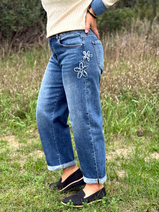 A person standing outdoors in a grassy field wearing blue denim jeans with white embroidered flower details on the back pockets, a cream ribbed sweater, and black slip-on shoes.