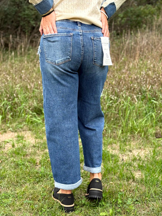 A person standing outdoors in a grassy field, shown from the back wearing blue jeans with a cream sweater and black shoes. A paper tag is attached to the back pocket of the jeans.