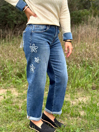 A person standing outdoors in a grassy field wearing blue jeans with three white embroidered starfish designs on the left leg, paired with a cream ribbed sweater and black slip-on shoes.