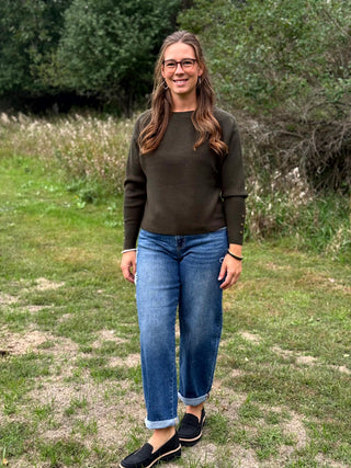 A woman wearing a dark olive green long-sleeve crewneck sweater, blue jeans, and black slip-on shoes, standing outdoors on a grassy field with trees in the background.