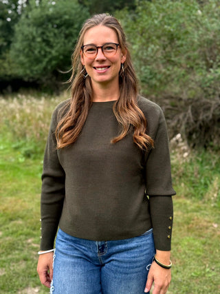 A woman wearing a dark olive green long-sleeve crewneck sweater, blue jeans, and black slip-on shoes, standing outdoors on a grassy field with trees in the background.