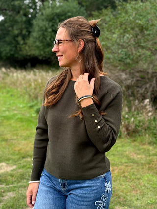 A woman wearing a dark olive green long-sleeve crewneck sweater, blue jeans, and black slip-on shoes, standing outdoors on a grassy field with trees in the background.