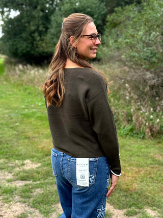 A woman wearing a dark olive green long-sleeve top, blue jeans with white embroidered details, and black slip-on shoes, standing outdoors on a grassy field with trees in the background.