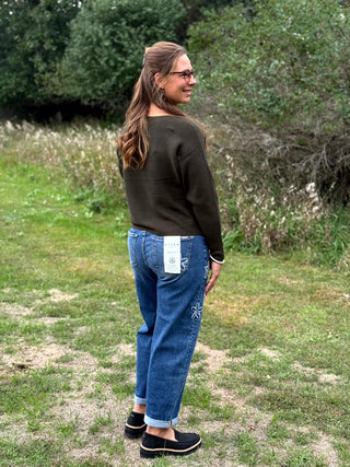 A woman wearing a dark olive green long-sleeve top, blue jeans with white embroidered details, and black slip-on shoes, standing outdoors on a grassy field with trees in the background.