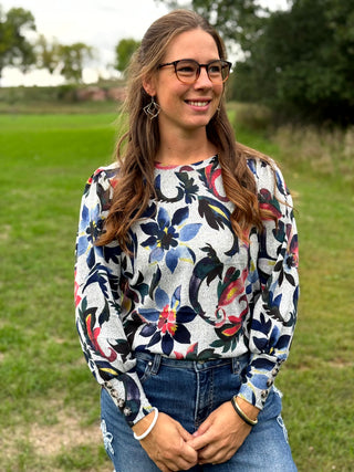 A woman with glasses and long brown hair stands outdoors in a grassy field, wearing a white long-sleeve top with a colorful floral pattern in blue, red, and green, paired with blue jeans.