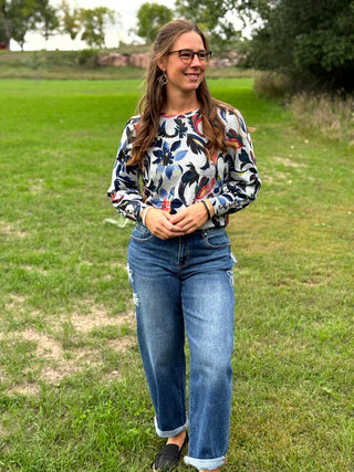 A woman wearing a long-sleeve floral print top and blue jeans stands outdoors on a grassy field.