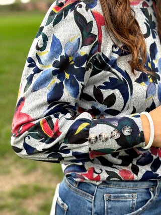 A close-up of a person wearing a gray long-sleeve knit top with a colorful floral pattern in blue, red, green, and yellow. The top features decorative buttons on the cuffs and is paired with blue jeans, shown outdoors with a blurred green background.