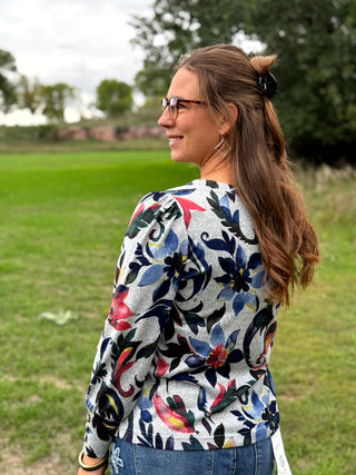 A woman standing outdoors on a grassy field, wearing a long-sleeve floral print top with blue jeans and black slip-on shoes.