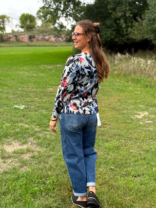A woman standing outdoors on a grassy field, wearing a long-sleeve floral print top with blue jeans and black slip-on shoes.