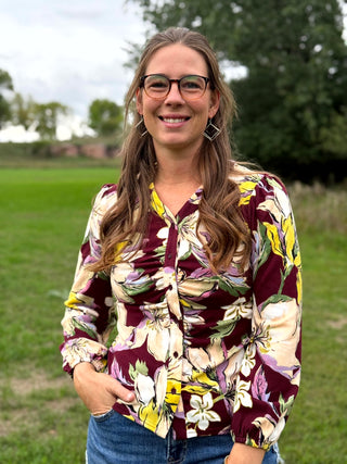 A woman wearing a floral long-sleeve blouse with a burgundy, yellow, and cream pattern, paired with blue jeans, standing outdoors in a grassy park.
