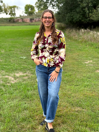 A woman wearing a floral long-sleeve blouse with a burgundy, yellow, and cream pattern, paired with blue jeans, standing outdoors in a grassy park.
