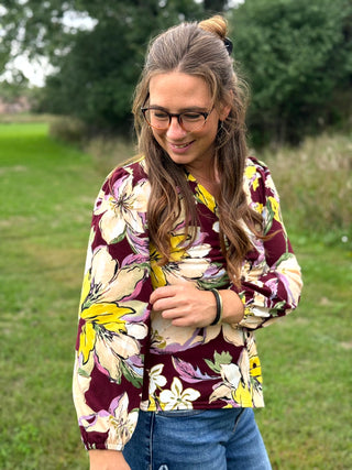 A woman wearing a floral long-sleeve blouse with a burgundy, yellow, and cream pattern, paired with blue jeans, standing outdoors in a grassy park.