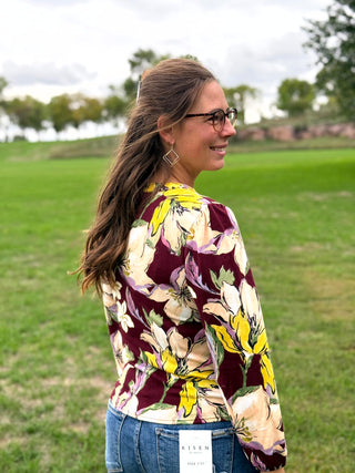 A woman wearing a floral long-sleeve blouse with a burgundy, yellow, and cream pattern, paired with blue jeans, standing outdoors in a grassy park.