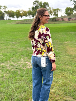 A woman wearing a floral long-sleeve blouse with a burgundy, yellow, and cream pattern, paired with blue jeans, standing outdoors in a grassy park.