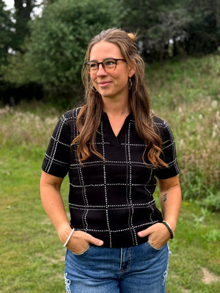 A woman with glasses and long brown hair stands outdoors in a grassy field, wearing a black short-sleeve knit top with a silver rhinestone windowpane grid pattern and blue jeans.