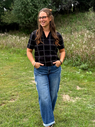 A woman wearing a black short-sleeve knit top with a silver rhinestone windowpane plaid pattern and blue jeans, standing outdoors on a grassy field with trees in the background.