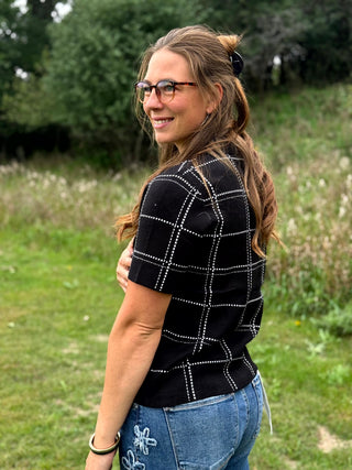 A woman with glasses and long brown hair stands outdoors in a grassy field, wearing a black short-sleeve knit top with a silver rhinestone windowpane grid pattern and blue jeans.