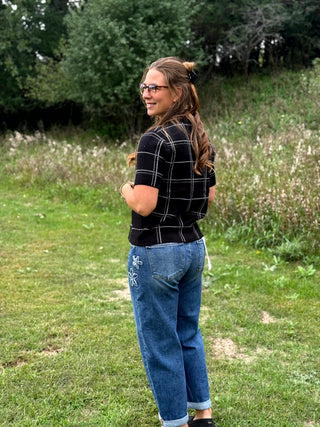 A woman with glasses and long brown hair stands outdoors in a grassy field, wearing a black short-sleeve knit top with a silver rhinestone windowpane grid pattern and blue jeans.