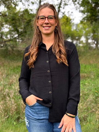 A woman with long brown hair and glasses stands outdoors in a grassy field, wearing a black long-sleeve button-up shirt and blue jeans.
