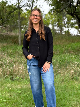 A woman with long brown hair and glasses stands outdoors in a grassy field, wearing a black long-sleeve button-up shirt and blue jeans.