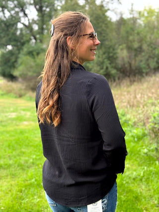 A woman wearing a black ribbed long-sleeve mock neck top and blue jeans stands outdoors in a grassy field with trees in the background.