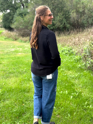 A woman wearing a black ribbed long-sleeve mock neck top and blue jeans stands outdoors in a grassy field with trees in the background.