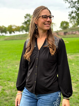 A woman wearing a black long-sleeve button-up blouse with patterned shoulder detailing and blue jeans, standing outdoors in a grassy park.